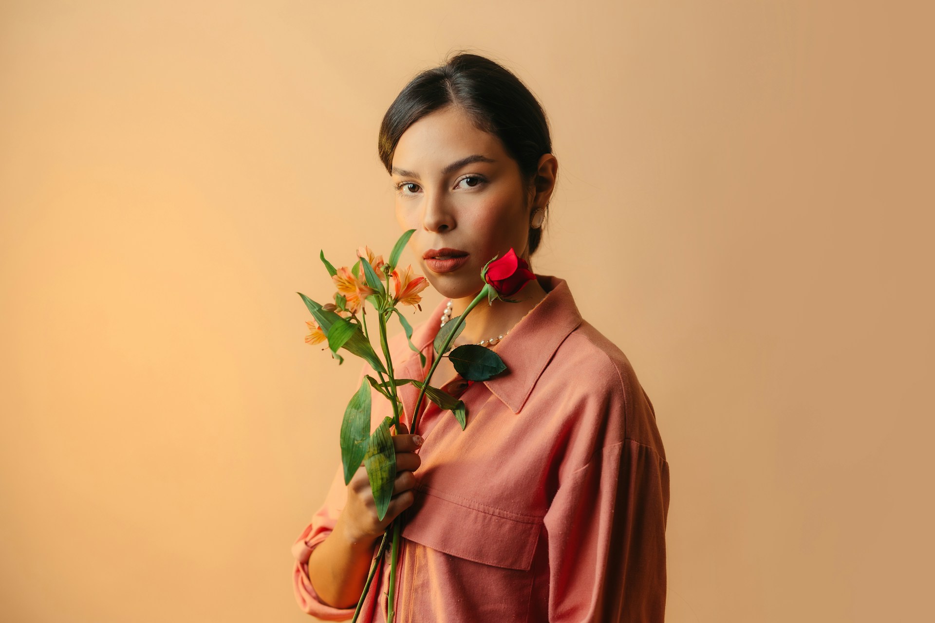 A woman in a pink shirt holding a red rose and orange flowers against a warm peach background.